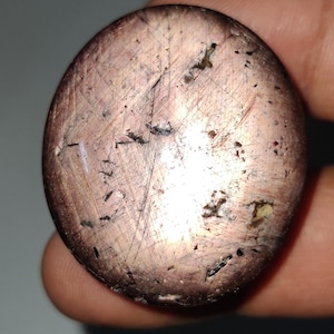 Könnte beinhalten: Ein runder, cabochon geschliffener Edelstein mit einem rosa und violetten Farbton. Der Stein hat einen hohen Glanz und ist mit kleinen, schwarzen Einschlüssen bedeckt.