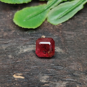 May include: A faceted, rectangular-cut, red gemstone. The gem is set against a dark, textured surface, with green leaves in the background. The gemstone's color is a vibrant, deep red, and it appears to be well-polished.
