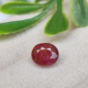 May include: A faceted oval-shaped gemstone with a deep red colour and visible dark inclusions throughout the stone. The gemstone features a polished surface with multiple triangular facets that reflect light across its surface.