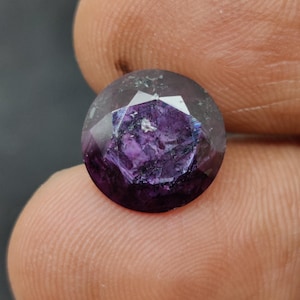 Puede incluir: Una gema redonda y facetada de color púrpura intenso. La gema tiene una apariencia clara y transparente con inclusiones internas y una superficie pulida. La piedra se sostiene entre los dedos, mostrando su tamaño y color.