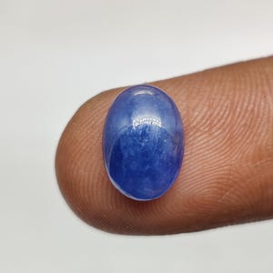 May include: An oval-shaped, cabochon-cut gemstone with a smooth, polished surface and a deep blue color. The stone exhibits a translucent quality with subtle internal variations and a reflective sheen across its rounded top surface.