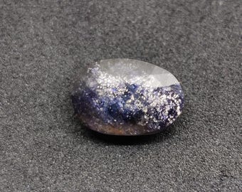 May include: Oval-shaped gemstone with a translucent, clear top and a base of deep blue and purple hues. The stone is speckled with silver-white inclusions, creating a galaxy-like effect. The polished surface reflects light.