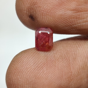 Könnte beinhalten: Ein rechteckiger, facettierter Edelstein in einem tiefen Rot-Rosa-Farbton. Der Stein hat eine klare, polierte Oberfläche mit sichtbaren inneren Einschlüssen und scharfen, kantigen Rändern, die eine lebendige Farbsättigung im Kristall zeigen.