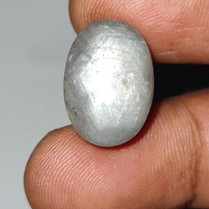May include: Oval-shaped, silvery-gray gemstone with a textured surface. The gem is held between two fingers, showcasing its polished appearance and natural color variations. The stone's shape and color suggest it could be a cabochon cut gemstone.