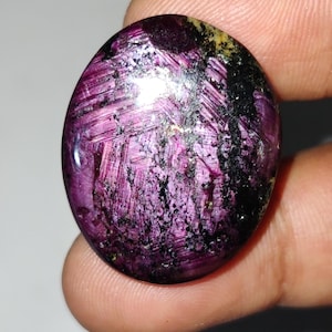 May include: A rounded, polished gemstone in a deep magenta colour, displaying a distinctive pattern of intersecting lines and dark, speckled inclusions. The stone's surface has a glossy finish, reflecting light and showing its unique texture.