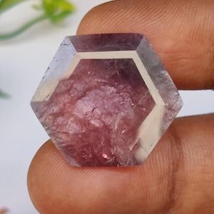 May include: A faceted, hexagonal gemstone with a translucent, pale purple exterior and a deep reddish-purple interior. The gem is held between fingers, showcasing its polished surfaces and unique color variations. The gem is a unique piece of jewelry.
