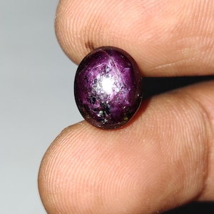 Peut inclure: Une pierre précieuse ronde et polie, de couleur violet foncé. La gemme présente un effet étoilé visible à sa surface, avec un centre brillant et réfléchissant. La pierre est tenue entre deux doigts, mettant en valeur sa taille et sa forme.