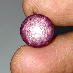 May include: A round, deep magenta gemstone with a star-like pattern. The gem is held between two fingers, showcasing its polished surface and internal light reflection. The gem's color is a rich, dark pink.