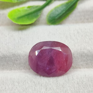 May include: An oval-shaped, faceted ruby gemstone with a deep pinkish-red hue. The stone displays a polished surface with multiple geometric facets that reflect light, showcasing its translucent quality and vibrant, saturated colour profile.