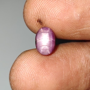 May include: An oval-shaped cabochon gemstone with a deep purple and magenta hue, featuring a distinct six-rayed star effect known as asterism that radiates from the center of the polished, smooth, and translucent surface of the stone.