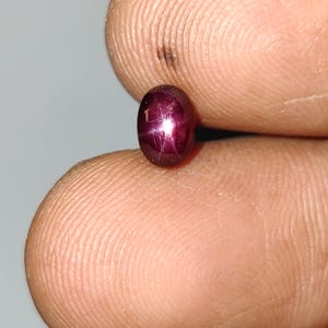 May include: A polished, oval ruby gemstone. The deep red gem has a star-like effect, with light radiating from the centre. The ruby is held between two fingers, showcasing its size and colour.