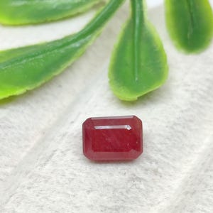 May include: A rectangular, faceted red gemstone with a polished surface and sharp, clean edges. The stone displays a deep, consistent crimson hue throughout its structure, showcasing a classic cut that reflects light across its flat top surface.