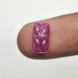 Op de afbeelding: Een rechthoekige, gesneden robijn edelsteen. De edelsteen heeft een dieproze kleur met een bloemmotief dat in het oppervlak is gesneden. De gravure is gedetailleerd en ingewikkeld, met een gestileerd bloemenpatroon.
