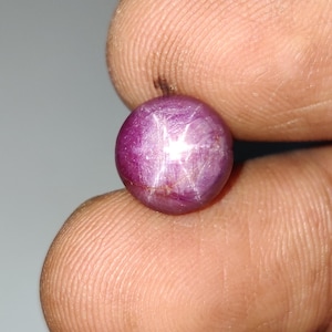 May include: A round, cabochon-cut star ruby gemstone with a deep magenta-red color. The stone displays a distinct six-rayed star effect, known as asterism, which appears as a bright white light reflection centered on the polished, smooth surface.