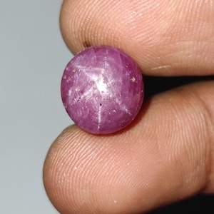 以下が含まれることがあります： 6条の星効果を持つ、丸く磨かれたルビーの宝石。宝石は濃いピンクがかった赤色で、指の間に挟まれているように見える。星の効果は、光を反射する白い星型の模様です。