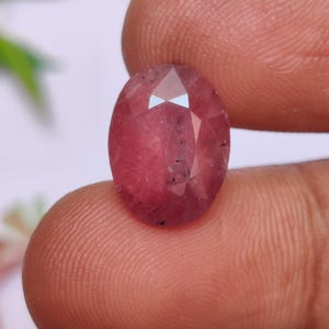 May include: A pinkish-red, oval-cut gemstone, held between fingers. The facets of the gem catch the light, highlighting its polished surfaces. The stone's colour varies, with darker inclusions visible within the gem.
