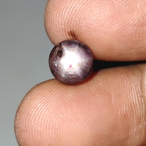 May include: A round, polished gemstone with a purplish-gray hue. The gem has a star-like effect, with a bright, reflective area in the center. The stone is held between two fingers, showcasing its size and shape. The gem's surface has subtle imperfections.