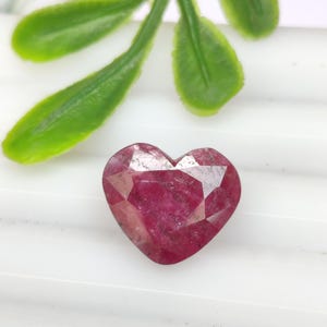 Op de afbeelding: Een hartvormige robijn edelsteen met een gefacetteerd oppervlak, met een diep magenta en rood kleurprofiel. De steen heeft een gepolijste afwerking met zichtbare interne insluitingen en een klassieke hartvormige slijpvorm met facetten.