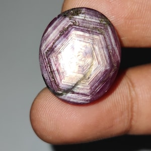 Op de afbeelding: Een gepolijste, ovaalvormige edelsteen met een stervormig patroon. De edelsteen heeft een dieppaarse tint met witte en lichtpaarse lijnen die vanuit het midden uitstralen. De steen wordt tussen de vingers gehouden, wat de grootte en vorm laat zien.