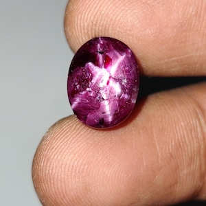 May include: An oval-shaped, deep magenta-colored gemstone with a cabochon cut. The stone displays a distinct six-rayed star effect across its polished, translucent surface, featuring internal crystalline inclusions and varied light reflections.