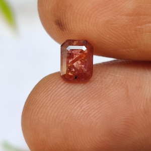 May include: A small, rectangular gemstone with a reddish-orange hue. The gem has a faceted cut, with visible internal inclusions. The stone is held between two fingers, showcasing its size and color.