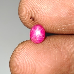 Puede incluir: Una pequeña piedra preciosa de color rosa vibrante y forma ovalada que presenta un patrón de estrella de seis puntas distintivo en su superficie lisa y pulida. La piedra muestra una calidad translúcida con tonos magenta profundos y un reflejo de luz brillante y centrado.