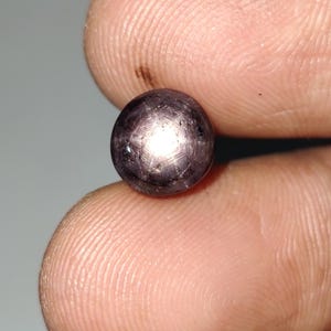 Op de afbeelding: Een donkerpaarse edelsteen met een ster-effect, vastgehouden tussen twee vingers. De steen heeft een ronde vorm en een gepolijst oppervlak dat licht reflecteert in een stervormig patroon. De kleur is diep, bijna zwart paars.