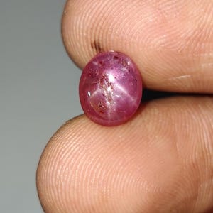 Puede incluir: Una gema rosa pulida, de forma ovalada, con un efecto de estrella. La gema tiene una apariencia translúcida con inclusiones internas y un sutil juego de luces. La gema se sostiene entre dos dedos.