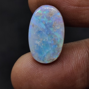 May include: An oval-shaped opal gemstone with a blue and iridescent color pattern. The opal has a polished surface and displays flashes of green and purple. The gemstone is held between two fingers against a dark background.