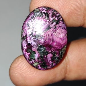 Puede incluir: Gema de forma ovalada con una vibrante mezcla de colores. La piedra presenta un tono magenta dominante, intercalado con inclusiones negras y verdes. La superficie tiene una estructura cristalina, que refleja la luz. Una pieza única y llamativa.