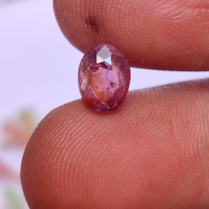 Puede incluir: Una gema ovalada facetada de color rosa-púrpura. La gema se sostiene entre dos dedos, mostrando sus superficies pulidas y variaciones de color internas. Las facetas de la gema reflejan la luz, creando un efecto brillante.