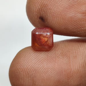 Può includere: Una pietra preziosa sfaccettata a taglio quadrato con una profonda tonalità rosso-arancio. La pietra presenta inclusioni interne e una qualità traslucida, con bordi geometrici netti e una superficie lucida che riflette la luce.