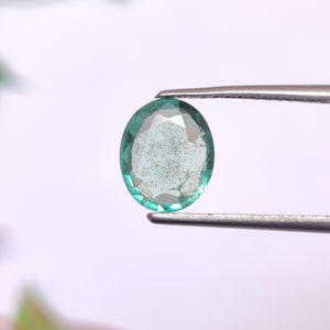 May include: A teal oval gemstone, held by tweezers, is shown against a white background. The gem has multiple facets and a slightly cloudy interior. The tweezers are silver-coloured.