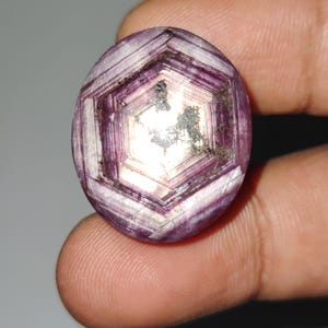 May include: Oval-shaped, faceted gemstone with a complex, geometric pattern. The gem features shades of purple and white, with a central, lighter-colored area. The stone's surface has a crystalline structure, and it appears to be held by a person's fingers.