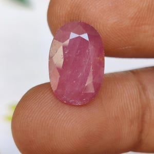 Könnte beinhalten: Ein facettierter, ovaler Rubin-Edelstein. Der Edelstein hat eine tiefrosa-rote Farbe und wird zwischen den Fingern gehalten. Der Schliff des Steins erzeugt mehrere reflektierende Oberflächen, die seine Brillanz verstärken. Der Edelstein ist ein natürlicher, unbehandelter Stein.