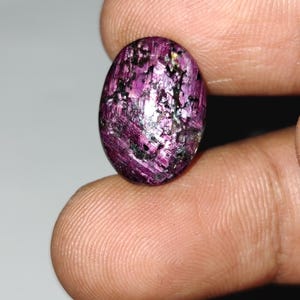 May include: An oval gemstone, polished to a high sheen, displaying a rich magenta colour with black and silver flecks. The gem is held between fingers, highlighting its smooth, reflective surface. The stone's distinctive pattern and colour variations are clearly visible.