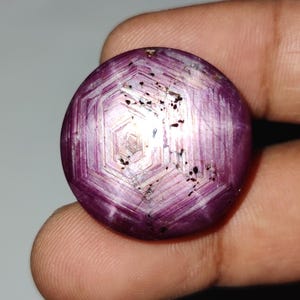 Pode incluir: Uma pedra preciosa redonda e polida com um tom magenta profundo. A gema apresenta um padrão semelhante a uma estrela e um design hexagonal em sua superfície. Pequenas inclusões pretas estão espalhadas pela pedra, adicionando à sua aparência única.