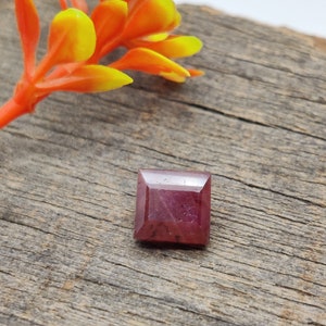 Puede incluir: Una piedra preciosa de color rojo intenso con forma cuadrada, sobre un fondo de madera rústico.