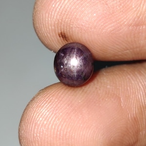 Pode incluir: Uma pequena pedra preciosa redonda, roxa escura, segurada entre os dedos. A gema tem um efeito de estrela, com linhas irradiando do centro. A pedra parece polida e lisa, refletindo a luz.
