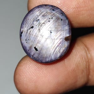 May include: Oval-shaped, polished gemstone with a lavender hue. The stone has a star-like effect with light reflecting off the surface. There are small black inclusions and a brown spot visible on the gem. The gemstone is held between fingers.