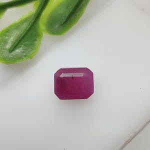 May include: A faceted, rectangular ruby gemstone with a deep red hue. The gem is set against a white background, with a sprig of green leaves in the upper left corner. The ruby's cut showcases its clarity and brilliance.