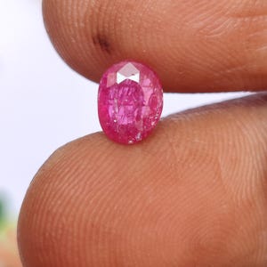 May include: A faceted, oval-shaped ruby gemstone. The gem is a vibrant pinkish-red color, with visible internal inclusions. The stone is held between two fingers, showcasing its size and clarity. This gemstone is suitable for jewelry making.
