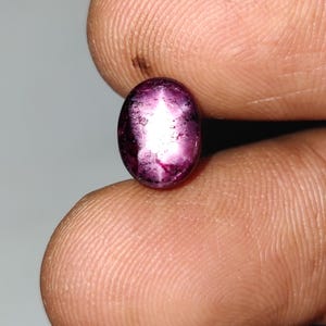 May include: Oval-shaped, deep red gemstone with a star-like pattern. The gem is held between fingers, showcasing its polished surface and color variations. This gemstone could be used in jewelry.