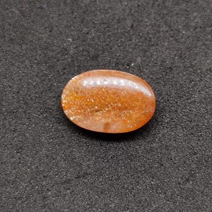 May include: Oval-shaped, polished gemstone with a warm orange hue and sparkling inclusions. The stone is smooth and rounded, with a translucent quality. The gemstone is set against a dark, textured background.