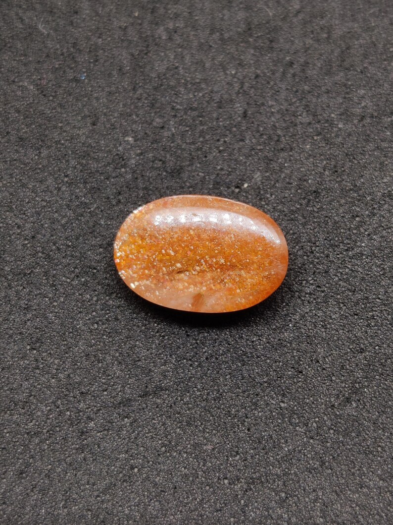 May include: Oval-shaped, polished gemstone with a warm orange hue and sparkling inclusions. The stone is smooth and rounded, with a translucent quality. The gemstone is set against a dark, textured background.