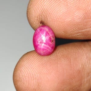 May include: A vibrant, oval-shaped cabochon star ruby gemstone displaying a distinct six-rayed star effect across its deep magenta-pink surface. The stone features a smooth, polished dome and internal inclusions that catch the light.