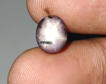May include: A small, oval-shaped, purplish gemstone with a star-like effect. The gem has a slightly cracked appearance with a dark green inclusion. The stone is held between two fingers against a neutral background.