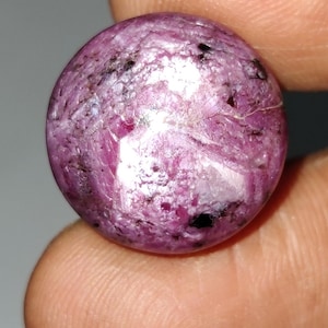 Puede incluir: Una gema redonda y pulida con un tono rosa intenso, salpicada de inclusiones negras y manchas blancas. La superficie de la gema refleja la luz, mostrando su estructura cristalina. Esta gema es un posible componente de joyería.
