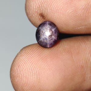 Może przedstawiać: Zbliżenie ciemnofioletowego kamienia szlachetnego z wzorem w kształcie gwiazdy. Owalny kamień ma wypolerowaną powierzchnię i jest trzymany między dwoma palcami. Klejnot ma właściwości odbijające światło.