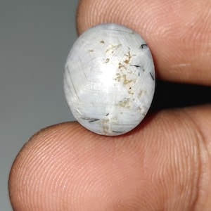 Op de afbeelding: Een ovaalvormige, gepolijste edelsteen met een doorschijnende, melkachtige witte basis. De steen heeft fijne zwarte en bruine insluitsels, waardoor een uniek patroon ontstaat. De edelsteen wordt tussen twee vingers gehouden.
